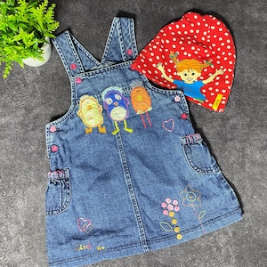 May include: A denim dungaree dress for a toddler, with embroidered cartoon birds and floral details. A red polka-dot hat with a cartoon girl graphic is included. The dress has pink button accents and pockets.