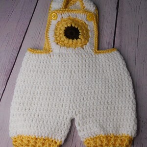 May include: White crocheted baby overalls with yellow trim and a sunflower design on the front.