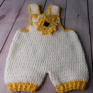 May include: White crocheted baby overalls with yellow trim and a crocheted sunflower on the front.