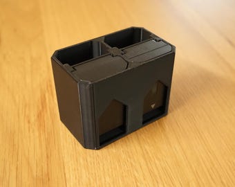 Peut inclure: Une station de charge de batterie noire, rectangulaire, imprimée en 3D en plastique, dotée de deux emplacements distincts. L'appareil a une finition mate et comprend deux compartiments internes avec des icônes de flèches pointant vers le bas.
