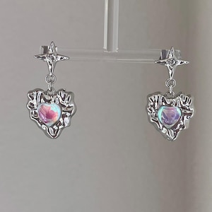 May include: A pair of silver earrings with a heart-shaped charm and a star charm. The heart charm has an iridescent finish and the star charm has a clear crystal.