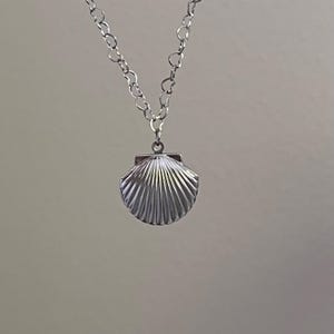 May include: A silver-colored necklace featuring a seashell pendant. The pendant has a detailed, fan-like design. The chain is made of small, interconnected links. The necklace is displayed against a neutral background, highlighting the jewelry's design.
