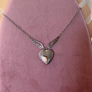 May include: A silver-colored heart-shaped locket necklace with wing accents. The locket has an ornate border and hangs from a delicate chain. The necklace is displayed on a pink surface.