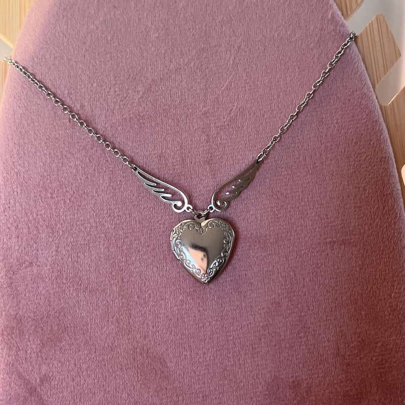 May include: A silver-colored heart-shaped locket necklace with wing accents. The locket has an ornate border and hangs from a delicate chain. The necklace is displayed on a pink surface.