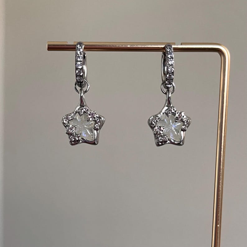 May include: A pair of silver hoop earrings with a dangling star charm. The star charm is made of clear crystal with a silver border and small silver accents.