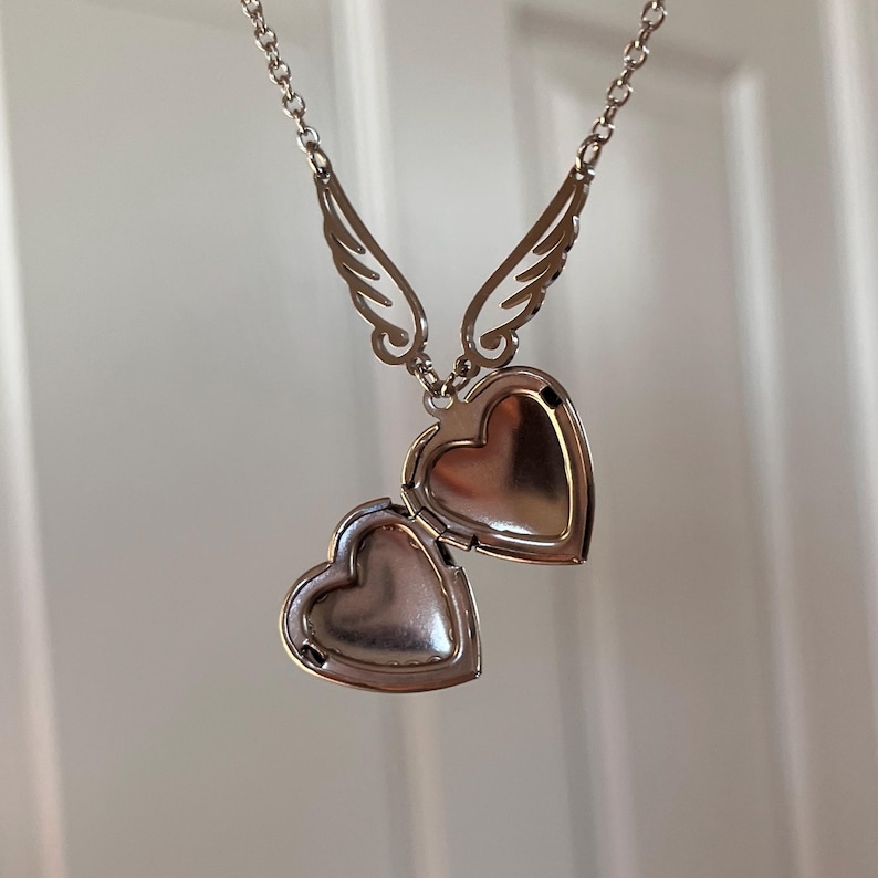 May include: Silver-toned heart-shaped locket necklace with openable compartment. The locket is suspended from a delicate chain and features decorative wing accents above the heart. The locket is open, revealing the interior space.