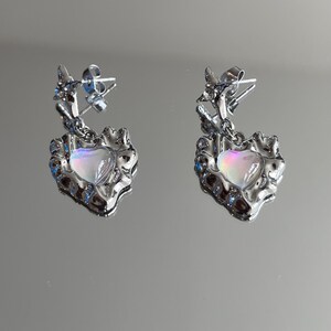 May include: A pair of silver earrings with a dangling heart-shaped charm. The heart charm has a iridescent, opalescent finish.