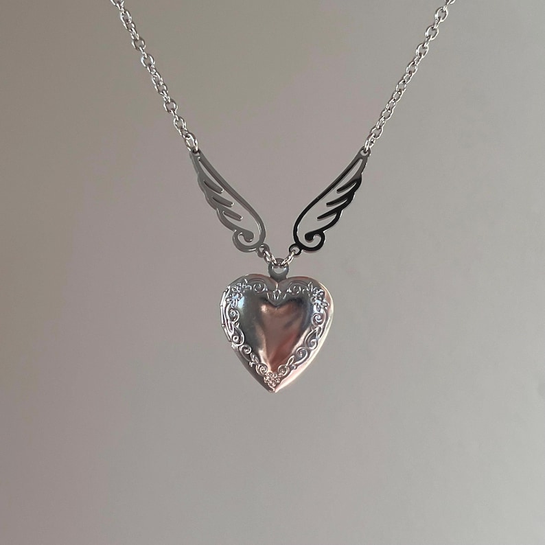 May include: A silver heart-shaped locket necklace with decorative edging and attached wing accents. The necklace hangs from a delicate chain, creating a romantic and elegant accessory. The locket is a classic piece of jewelry.