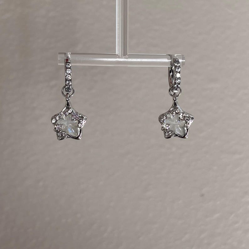 May include: A pair of silver star-shaped earrings with clear crystals. The earrings are hanging from a silver hoop.