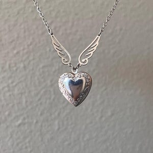 May include: A silver-colored necklace featuring a heart-shaped locket pendant with floral detailing. The pendant is suspended from a chain and flanked by stylized wing accents. The necklace is displayed against a neutral background.