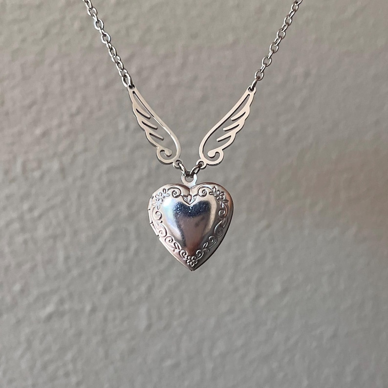 May include: A silver-colored necklace featuring a heart-shaped locket pendant with floral detailing. The pendant is suspended from a chain and flanked by stylized wing accents. The necklace is displayed against a neutral background.