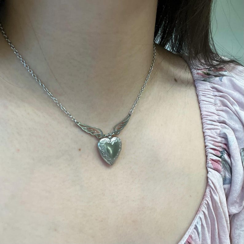 May include: Silver-toned heart-shaped locket necklace with delicate wing accents. The locket hangs from a fine chain, creating a romantic and elegant accessory. The heart pendant has a textured surface, adding detail to the design.