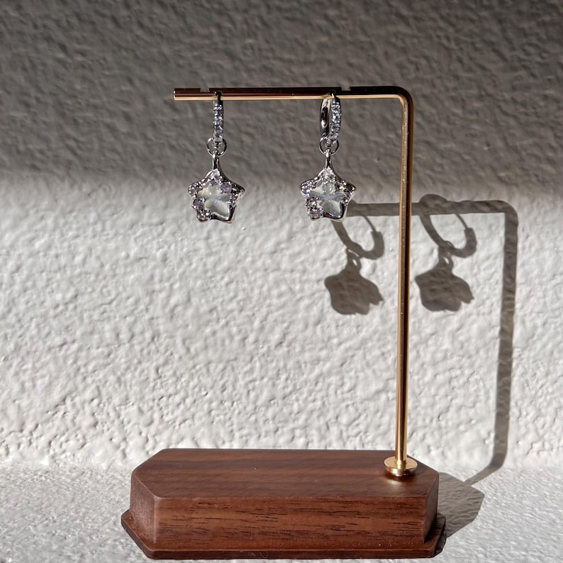 May include: A pair of silver star-shaped earrings with clear stones hanging from a gold stand.