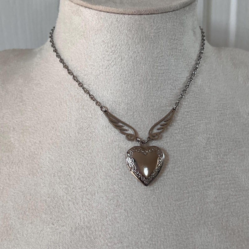 May include: A silver-toned necklace with a heart-shaped pendant. The pendant has an ornate, engraved design and is suspended from a chain. The necklace features small wing accents on either side of the heart.