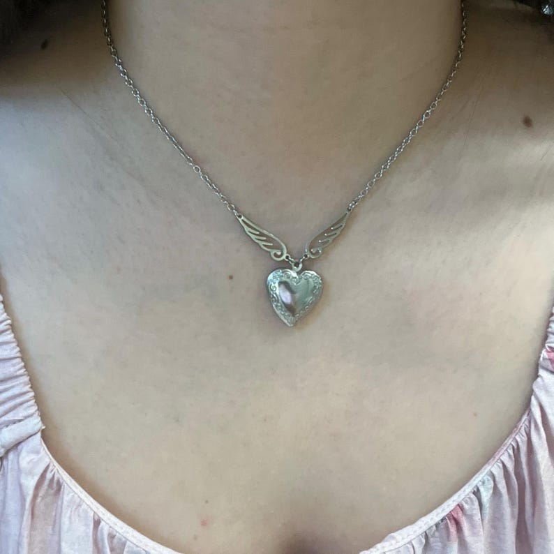 May include: Silver-toned necklace featuring a heart-shaped locket pendant with wing accents. The necklace is worn around the neck, with the pendant resting on the chest. The locket has a detailed, textured surface and hangs from a delicate chain.