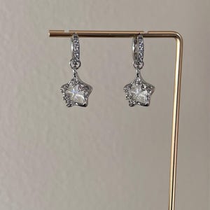 May include: A pair of silver hoop earrings with a dangling star charm. Each star charm is adorned with clear rhinestones.