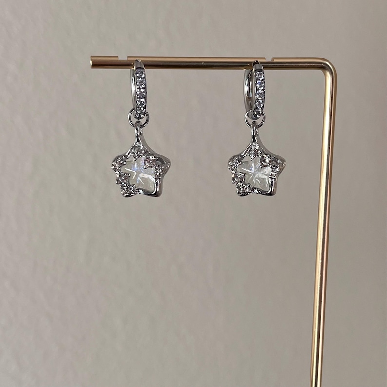 May include: A pair of silver hoop earrings with a dangling star charm. Each star charm is adorned with clear rhinestones.