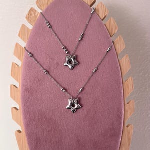 Peut inclure: Deux colliers en argent avec des pendentifs en forme d'&eacute;toile sur un pr&eacute;sentoir en velours rose. Les &eacute;toiles ont un trou au centre.