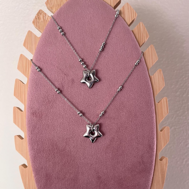 Peut inclure: Deux colliers en argent avec des pendentifs en forme d'&eacute;toile sur un pr&eacute;sentoir en velours rose. Les &eacute;toiles ont un trou au centre.