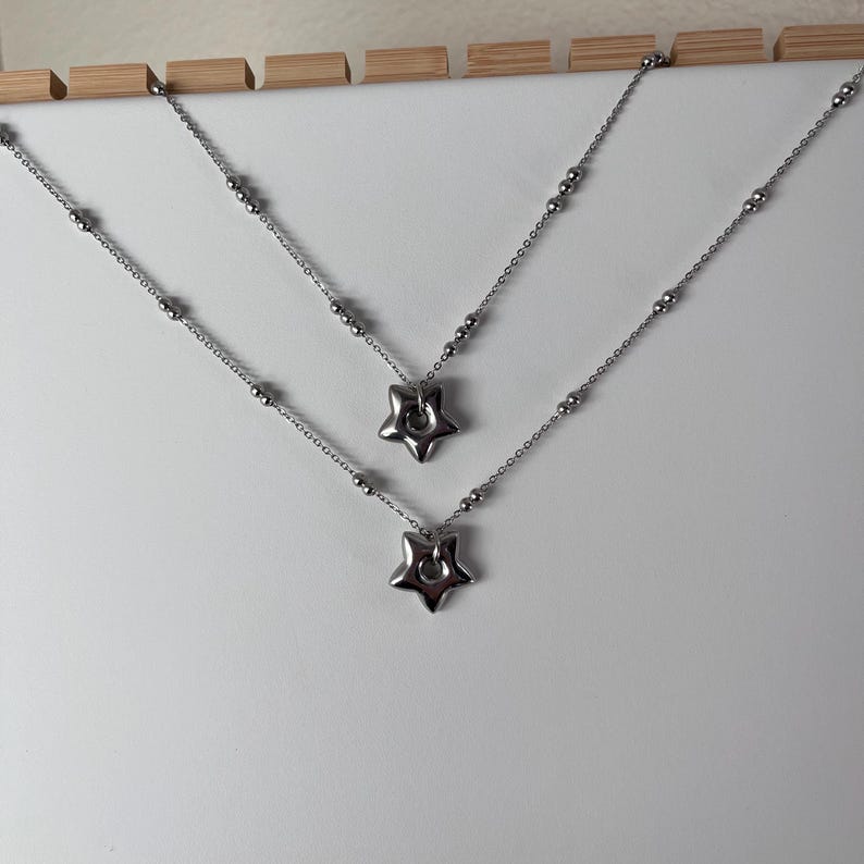 Peut inclure: Deux colliers en argent avec des pendentifs en forme d'&eacute;toile. Les colliers sont superpos&eacute;s sur un fond blanc.