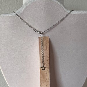May include: A silver-colored necklace with a delicate chain and a small star pendant. The necklace is displayed on a wooden stand against a white background. The star pendant hangs from an adjustable chain.