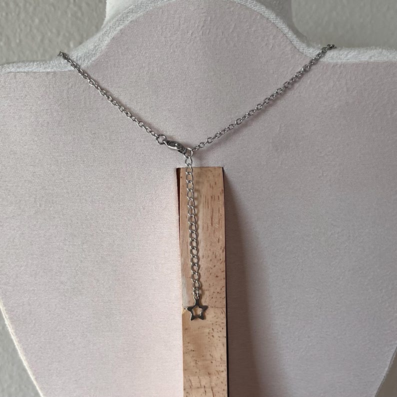 May include: A silver-colored necklace with a delicate chain and a small star pendant. The necklace is displayed on a wooden stand against a white background. The star pendant hangs from an adjustable chain.
