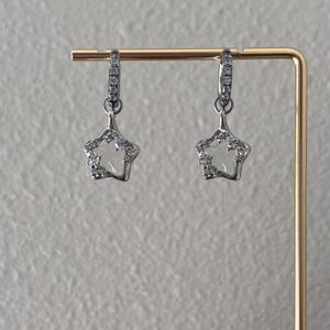 May include: A pair of silver star-shaped earrings with clear crystal accents. The earrings are hanging from a gold display stand.