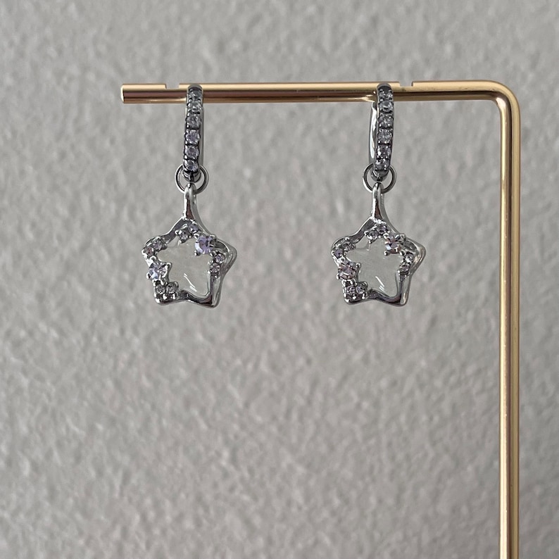 May include: A pair of silver star-shaped earrings with clear crystal accents. The earrings are hanging from a gold display stand.