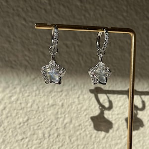 May include: A pair of silver hoop earrings with a dangling star charm. The star charm is adorned with clear rhinestones.