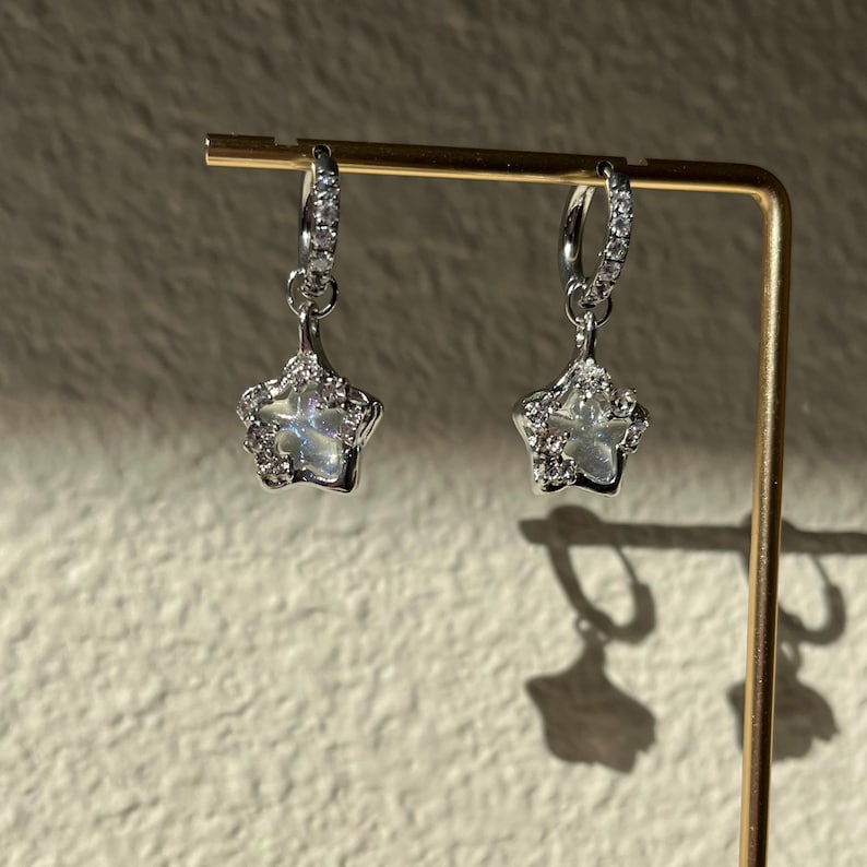 May include: A pair of silver hoop earrings with a dangling star charm. The star charm is adorned with clear rhinestones.