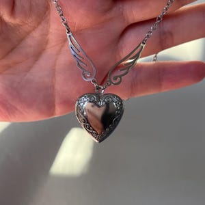 May include: A silver-colored heart-shaped locket necklace with decorative floral detailing around the edges. The necklace features delicate wing-shaped accents on either side of the chain, adding a whimsical touch to the jewelry.