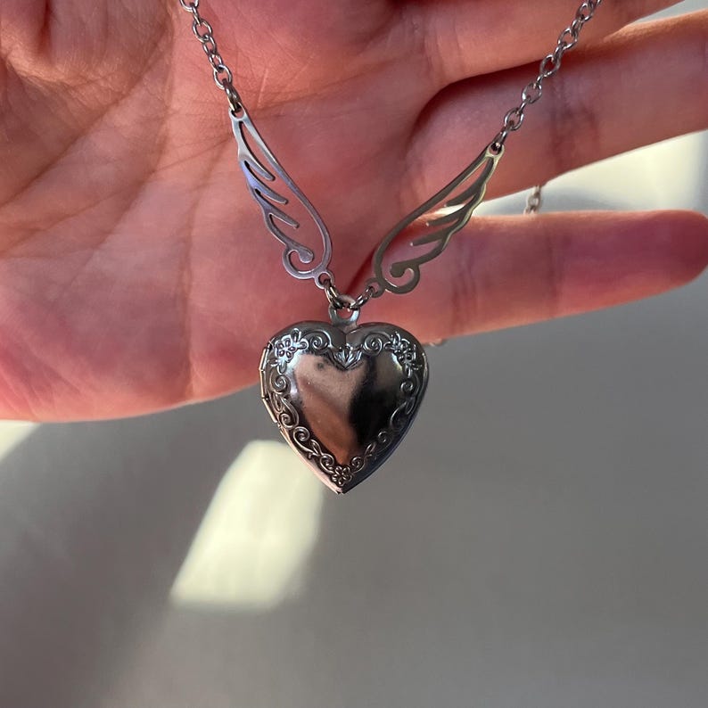May include: A silver-colored heart-shaped locket necklace with decorative floral detailing around the edges. The necklace features delicate wing-shaped accents on either side of the chain, adding a whimsical touch to the jewelry.