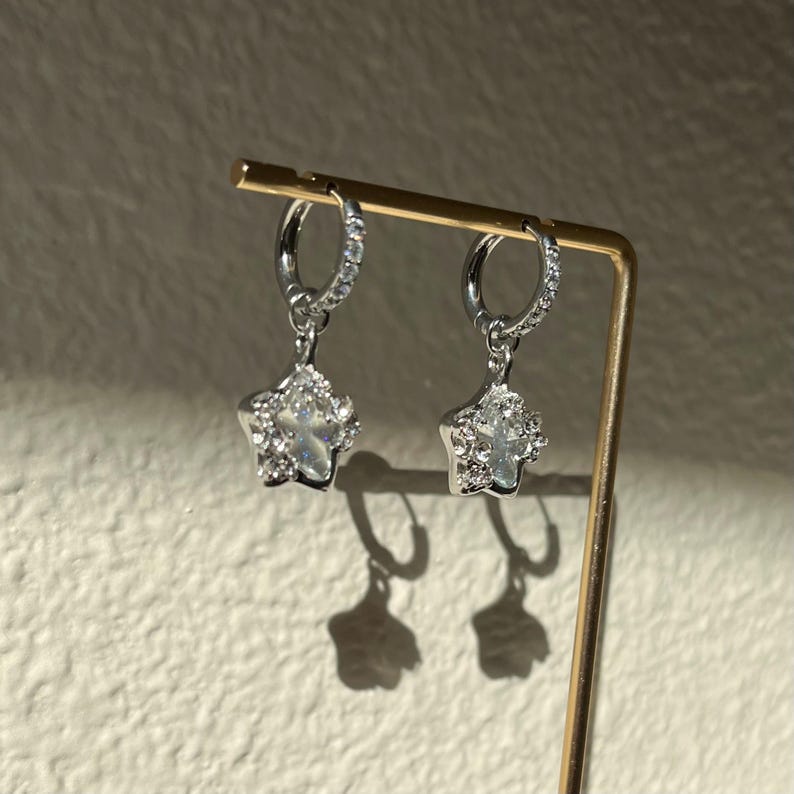 May include: A pair of silver hoop earrings with a dangling star charm. Each star charm is encrusted with clear cubic zirconia stones.