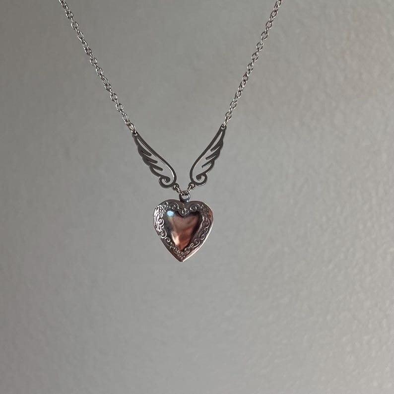 May include: A silver-toned necklace featuring a heart-shaped locket pendant with an ornate design. The pendant is suspended from a chain and flanked by stylized wing accents. The necklace is displayed against a neutral background.