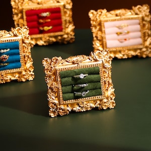 May include: Ornate gold-colored picture frames with velvet ring holders in various colors: red, blue, green, and pink. Each frame displays multiple rings. The frames are arranged on a dark green surface, showcasing the jewelry.