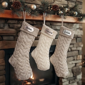 Peut inclure: Trois bas de Noël en tricot torsadé aux tons neutres sont suspendus à une cheminée en bois. Chaque bas a une étiquette métallique personnalisée avec un nom : "Nolan", "Fannie" et "Uriah". Les bas sont décorés d'une guirlande festive.