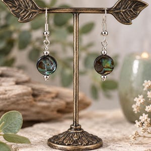 May include: A pair of drop earrings featuring round, iridescent abalone shell beads with dark green and blue tones. The beads are accented with small, polished silver-tone metal spacer beads and suspended from silver-tone metal fishhook ear wires.