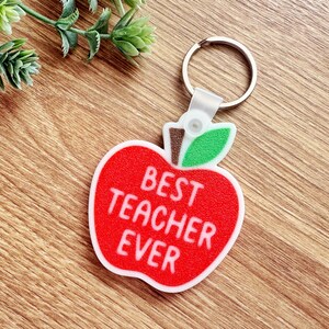Könnte beinhalten: Ein roter, apfelförmiger Schlüsselanhänger mit den weißen Worten "BEST TEACHER EVER". Der Schlüsselanhänger hat ein grünes Blatt und einen braunen Stiel. Der Schlüsselanhänger ist an einem silbernen Schlüsselring befestigt.