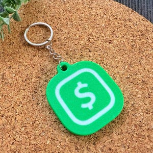 May include: A green keychain with a white dollar sign inside a white square. The keychain is attached to a silver key ring.