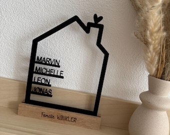 May include: A black house-shaped family sign with the names MARVIN, MICHELLE, LEON, and JONAS, and the text Familie WINKLER. The sign is mounted on a wooden base and sits on a wooden shelf. A beige vase with dried flowers is to the right.