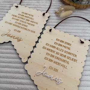 Könnte beinhalten: Zwei Holzschilder mit gewellten Kanten und braunen Aufhängeschnüren. Die Schilder zeigen deutschen Text und den Namen "Jonas" in Schreibschrift. Ein Schild hat den Text "Ich bin mutig" und das andere sagt "Ich bin stark".