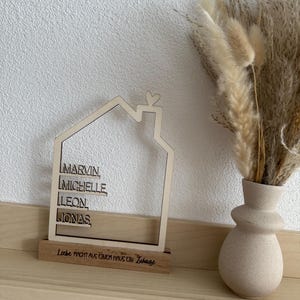 May include: A wooden house-shaped decoration with the names MARVIN, MICHELLE, LEON, and JONAS. The base reads "Liebe macht aus einem Haus ein Zuhause." A beige vase with dried flowers sits to the right.