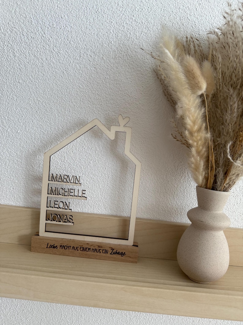 May include: A wooden house-shaped decoration with the names MARVIN, MICHELLE, LEON, and JONAS. The base reads "Liebe macht aus einem Haus ein Zuhause." A beige vase with dried flowers sits to the right.