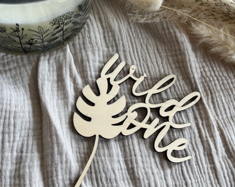 May include: A wooden cake topper with a leaf design and the words "wild one" in a cursive font. The topper is on a light brown fabric background.