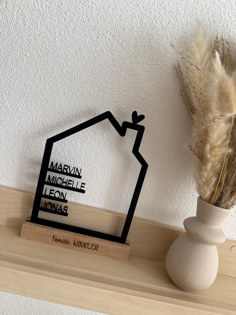 May include: A black house-shaped family sign with the names Marvin, Michelle, Leon, and Jonas. The sign sits on a wooden base with the text "Familie Winkler". A small heart is at the top of the house outline.