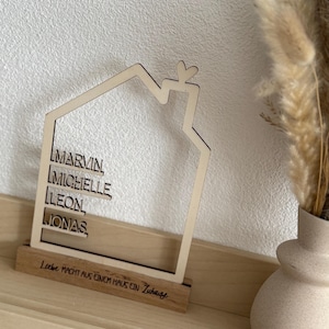 May include: A wooden house-shaped decoration with the names MARVIN, MICHELLE, LEON, and JONAS. The house has a heart-shaped detail at the top and sits on a wooden base with the text "Liebe macht aus einem Haus ein Zuhause."