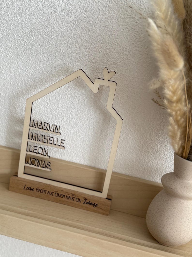 May include: A wooden house-shaped decoration with the names MARVIN, MICHELLE, LEON, and JONAS. The house has a heart-shaped detail at the top and sits on a wooden base with the text "Liebe macht aus einem Haus ein Zuhause."