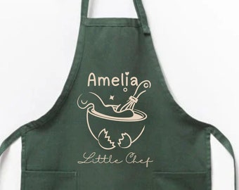 May include: Green apron with a white design of a mixing bowl, whisk, and eggs. The text "Amelia" and "Little Chef" is printed on the apron.