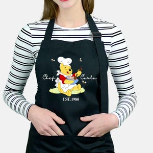 May include: A black apron featuring a graphic of Winnie the Pooh wearing a white chef hat and apron, holding a blue bowl and spoon. The text reads Chef Carla and EST. 1980, surrounded by small bees. The apron has a front pocket and neck strap.