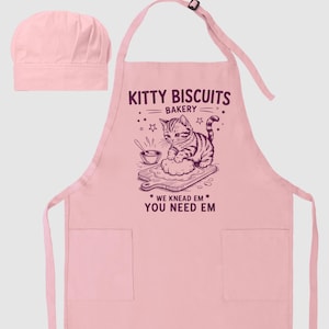 May include: A pink apron and chef's hat set. The apron features the text "Kitty Biscuits Bakery" with an illustration of a cat kneading dough. The text "We Knead Em, You Need Em" is also on the apron.